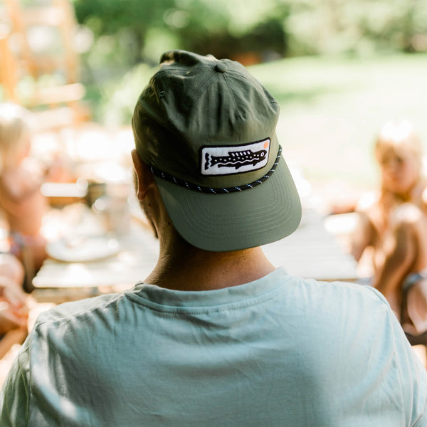Trout Hat worn by a person outdoors, showcasing its adjustable design and unique trout logo in a casual setting.