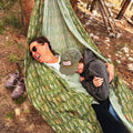Two people relaxing in a Double Hammock & Pro Hanging Straps (River Trout Print) in a forest setting.