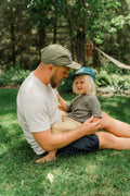 Dad in an olive Trout Hat and a child in a blue Trout Hat sitting together on backyard grass, Trek Light