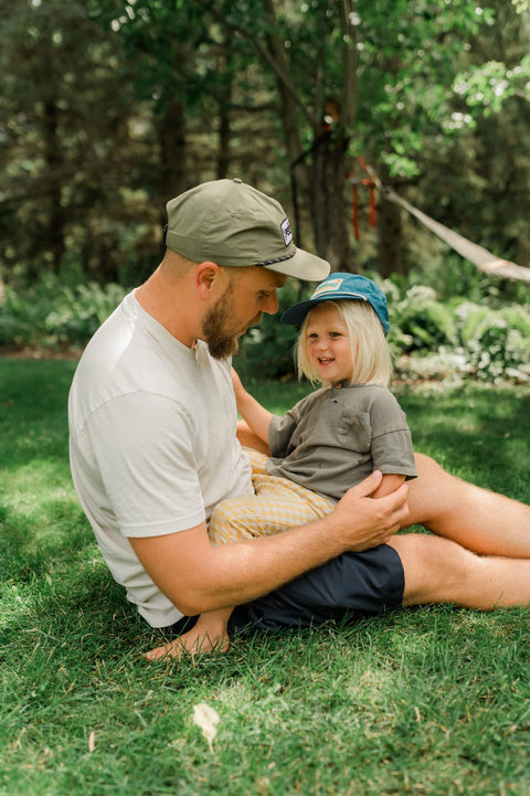 Dad in an olive Trout Hat and a child in a blue Trout Hat sitting together on backyard grass, Trek Light