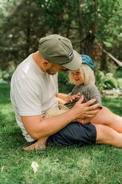 Man in an olive Trout Hat leaning toward a child in a blue Trout Hat on a green lawn, Trek Light