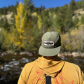 Person wearing a Trout Hat by a serene river with autumn trees in the background.