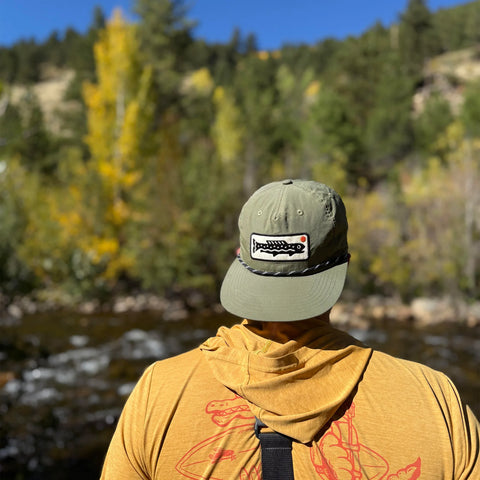 Person wearing a Trout Hat by a serene river with autumn trees in the background.