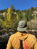 Person wearing a Trout Hat by a serene river with autumn trees in the background.