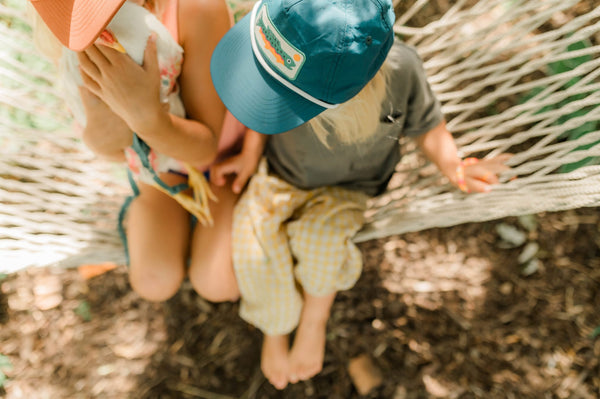 Toddler wearing a Trout Kids Hat while sitting on a hammock with a sibling, enjoying outdoor fun.