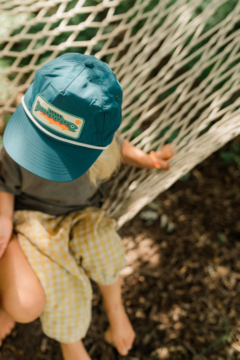 Child wearing a Trout Hat Twinsie Set sitting in a hammock, enjoying a sunny outdoor moment.