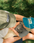Child in a teal Trout Kids Hat with a green trout patch sits on grass in an adult's lap, seen from above.