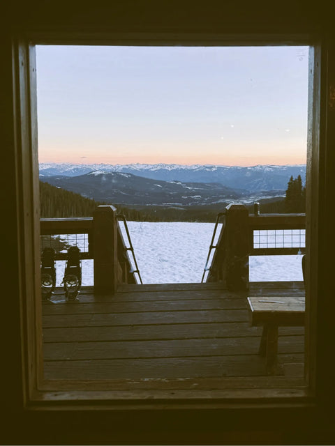Scenic view of snow-covered mountains and sunset through a rustic window frame at a mountain cabin.