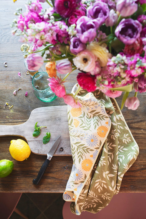 Wavy Flowers Kitchen & Tea Towel displayed on a wooden table with fresh lemons, limes, and a floral arrangement.