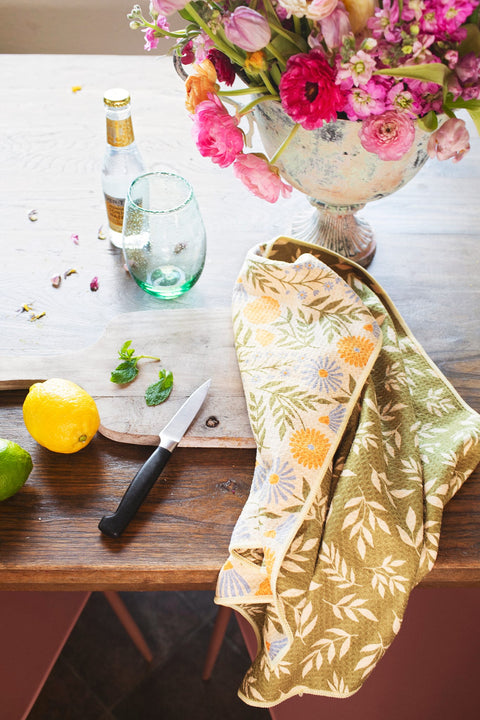 Wavy Flowers Kitchen & Tea Towel on a wooden table with fruits, flowers, and a glass, showcasing its floral design.