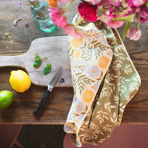 Wavy Flowers Kitchen & Tea Towel draped over a wooden board with lemons, limes, and flowers in a vase.