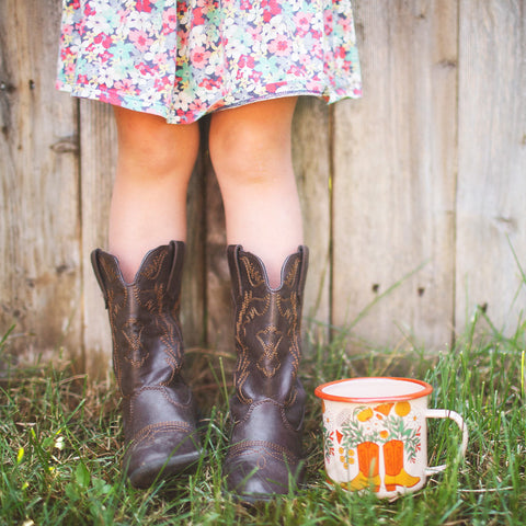 Western Bloom Enamel Mug 16oz by Trek Light with cowboy boots and floral dress in a rustic outdoor setting.