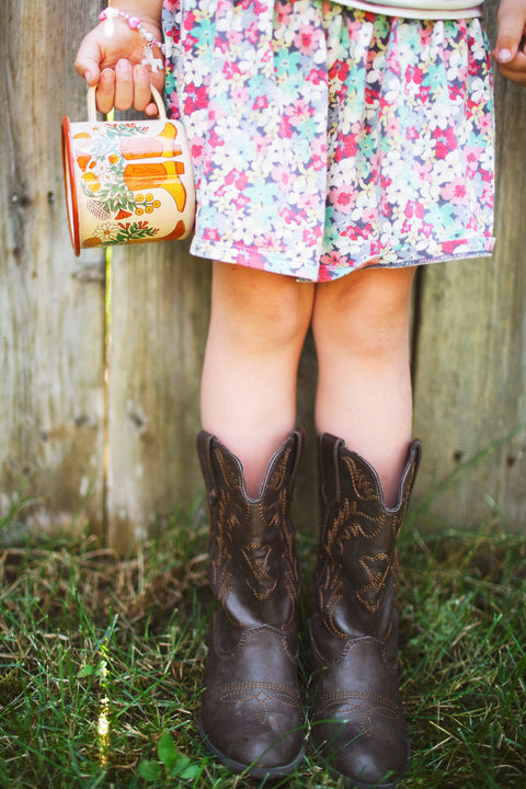 Western Bloom Enamel Mug 16oz in a child's hand, paired with brown cowboy boots and a floral dress.