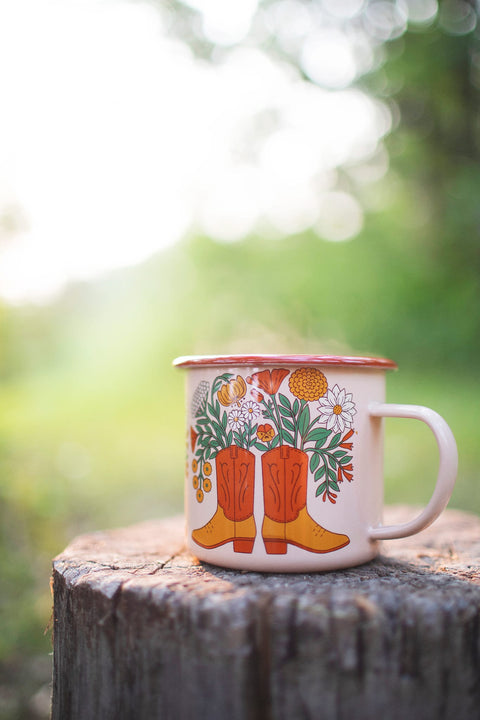 Western Bloom Enamel Mug 16oz with floral and cowboy boot design on a wooden surface outdoors.