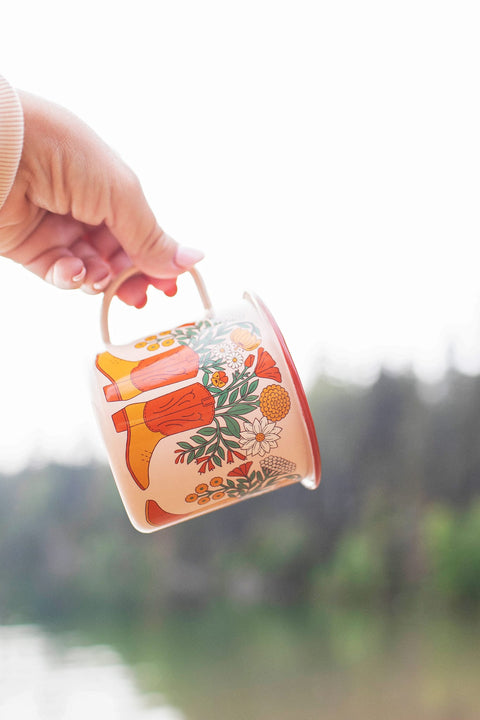 Western Bloom Enamel Mug 16oz being held outdoors, showcasing vibrant floral and cowboy boot design.