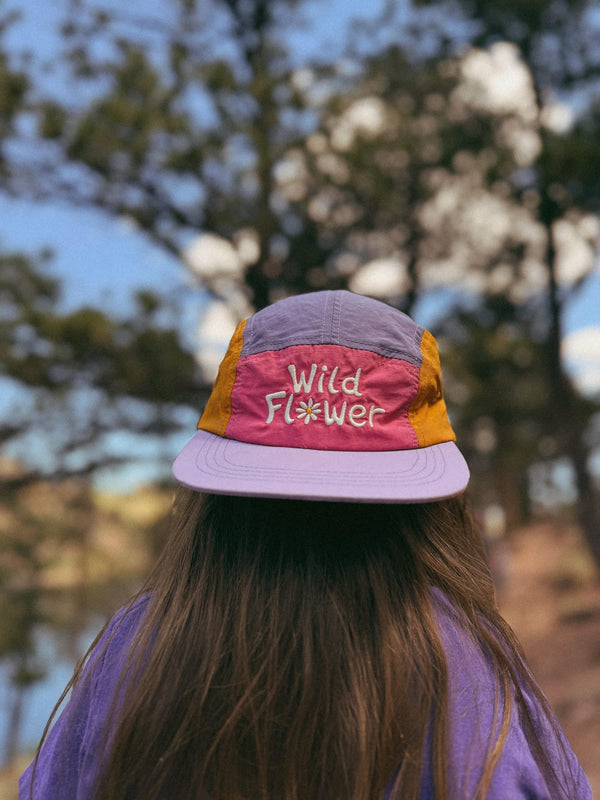 Child wearing a colorful hat with the text 'Wild Flower Kids Hat' against a natural backdrop.