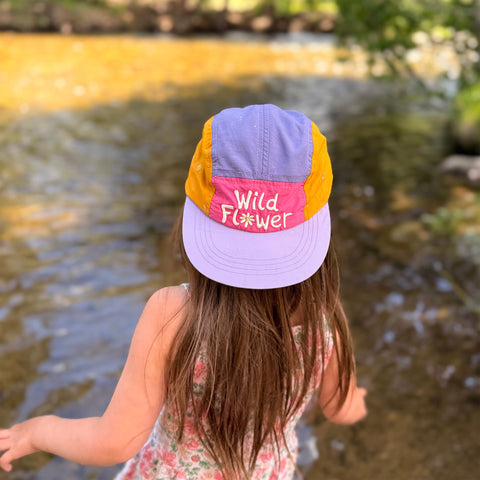 Child wearing a colorful Wild Flower Kids Hat by a river, highlighting style and fun for outdoor adventures.