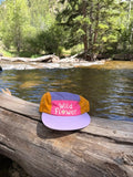 Wild Flower Kids Hat placed on a log by a peaceful stream surrounded by trees.