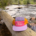 Wild Flower Kids Hat resting on a log by a stream, showcasing vibrant colors and playful design for adventures.