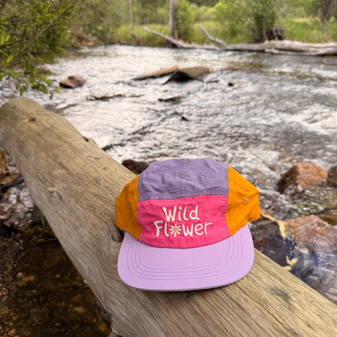 Wild Flower Kids Hat resting on a log by a stream, showcasing vibrant colors and playful design for adventures.