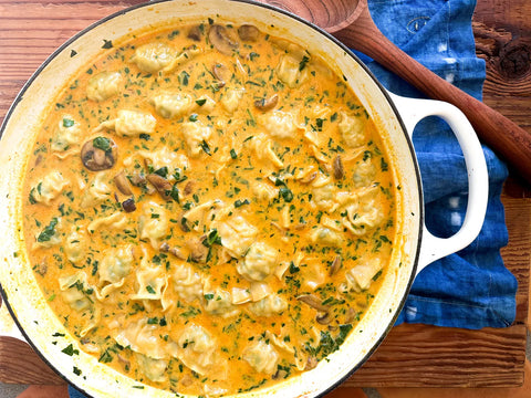 Creamy mushroom ravioli in a white skillet on a wooden table with blue napkin, garnished with fresh herbs.