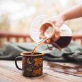 Pouring coffee into a 16oz Woodland Friends enamel mug by Trek Light, featuring adorable forest animals design, on a wooden table.