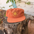 You Are My Sunshine Hat displayed on a rustic wooden stump, featuring a vibrant orange color and cheerful design.