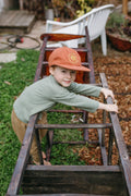 Child wearing Trek Light 'You Are My Sunshine' hat, playing outdoors with a rustic wooden frame, embodying adventure and joy.