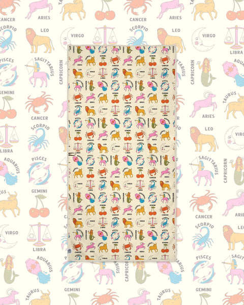 Zodiac Wander & Travel Towel in cream with colorful animal and sign illustrations, laid flat on a matching pastel pattern.