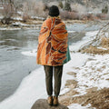Person standing by a snowy river wrapped in an Alpenglow Wander Towel with earthy tones. Part of Trek Light’s sustainable line.