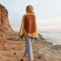 Woman wrapped in Trek Light blanket standing by the seaside cliffs during sunset.