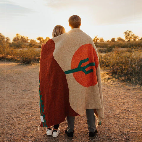 Couple wrapped in a cozy handwoven Arizona Blanket, made from recycled materials, enjoying a sunset in the desert. By Trek Light.