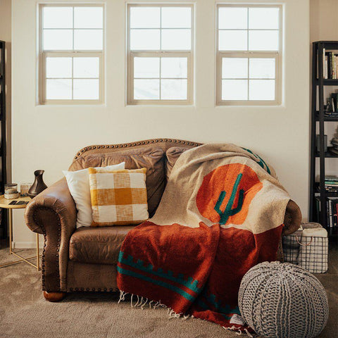 Cozy Trek Light Arizona Blanket on a couch, handwoven from recycled materials, featuring a desert sunset and cactus design. Beautiful and versatile.