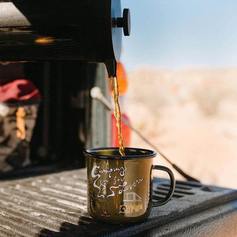 Pouring coffee into a 'Camping Is For Lovers' enamel mug by Trek Light, perfect for campfire adventures and cozy sips.