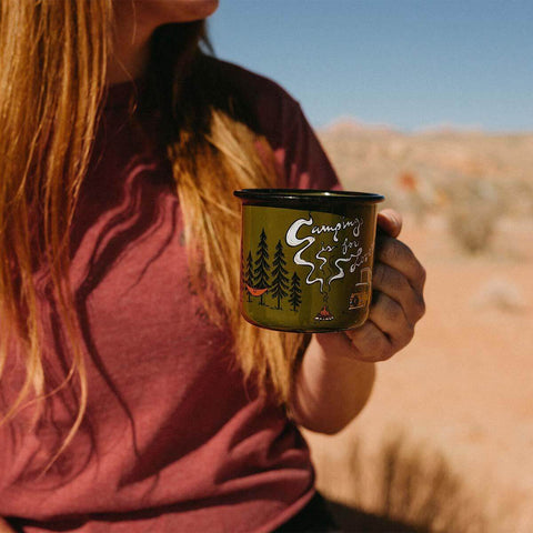 Person holding a 12oz Trek Light enamel mug with 'Camping Is For Lovers' graphic, perfect for outdoor adventures and cozy moments.