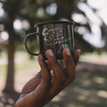 Hand holding Trek Light 'Camping Is For Lovers' enamel mug in nature, emphasizing adventure and durability.