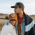 Couple wrapped in Trek Light blanket outdoors, both wearing hats, enjoying a cozy moment in nature.