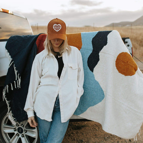 Woman wearing a brown 'Camping Is For Lovers' hat by Trek Light, leaning on colorful blanket with open landscape backdrop.