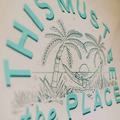 Close-up of "This Must Be The Place" canvas flag with tropical design, featuring palm trees and a relaxing hammock. Trek Light product.