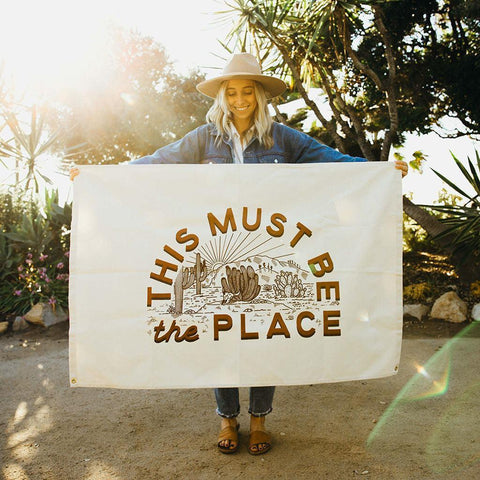 Woman holding Trek Light 'This Must Be The Place' canvas flag made of natural cotton, perfect for home decor and adventure lovers.