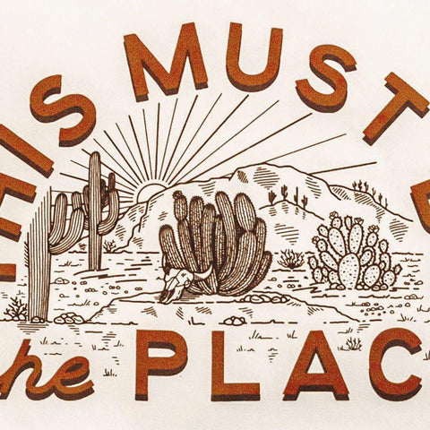 "This Must Be The Place" canvas flag with desert illustration, featuring cacti and sunburst, made by Trek Light. Cozy decor with brass grommets.