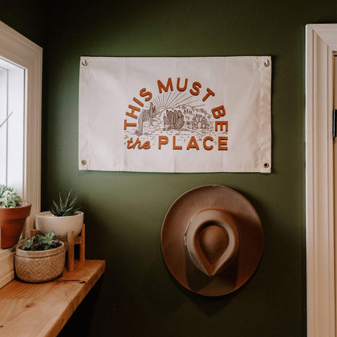 This Must Be The Place canvas flag on green wall, featuring hand-screened print. Trek Light artisanal decor with brass grommets.