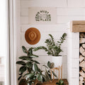 Trek Light 'This Must Be The Place' canvas flag on white wall, accompanied by green plants and a stylish hat.