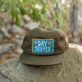 Vintage-style Day Tripper hat by Trek Light, 6-panel snapback with a flat brim, resting on a rock in a natural setting.