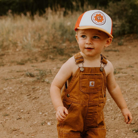 Toddler wearing a Trek Light Earth Day Kids Hat, enjoying an outdoor adventure in brown overalls.