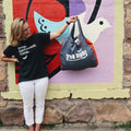 Woman smiling with Trek Light Eco Tote Deluxe in front of colorful mural, promoting eco-friendly, plastic-free living.