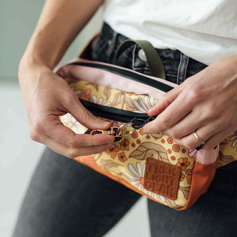 Person wearing Trek Light Everyday Fanny Pack (Bloom), adjusting the zipper. Converts to hip pack or shoulder sling for versatile style.