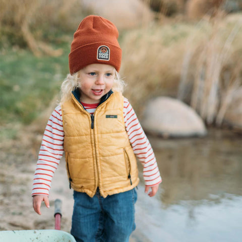 Child near water wearing a Good Vibes Kids Beanie by Trek Light, spreading positivity and charm in a cozy outdoor setting.