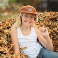 Child wearing Good Vibes Sherpa Kids Hat by Trek Light, sitting in autumn leaves. Cozy style with corduroy and sherpa flaps. Fun outdoors.