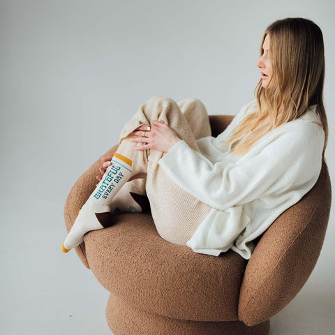 Woman relaxing in brown chair wearing cozy 'Grateful Every Day' Trek Light socks, perfect for comfort and style.